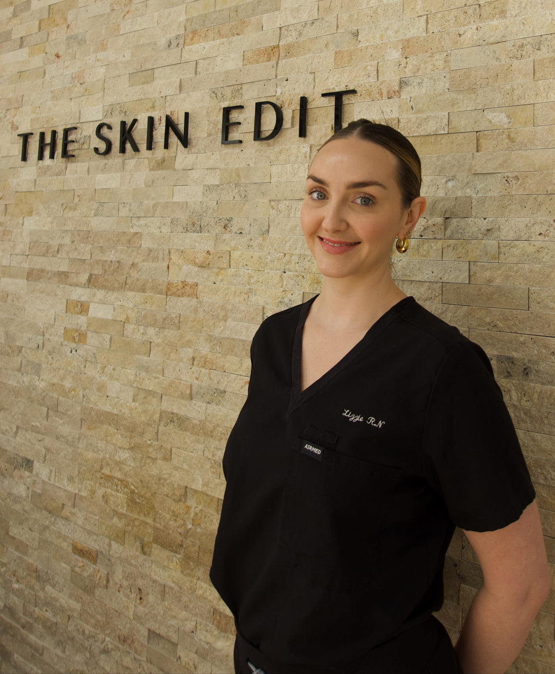 Woman in a black scrub top standing in front of a stone wall with 'The Skin Edit' sign.