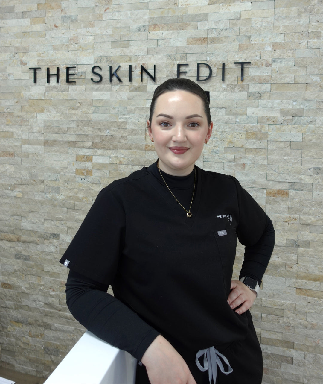 Person wearing a black outfit with 'The Skin Edit' logo in front of a textured wall.